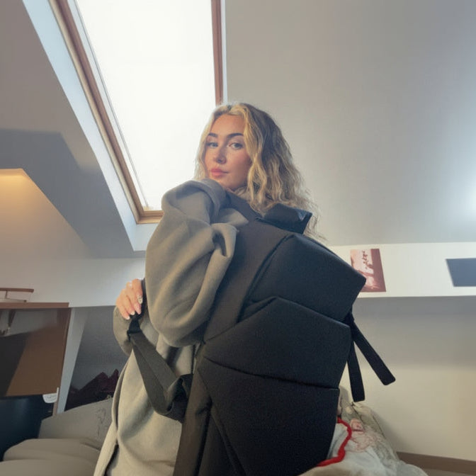 Person holding a large black backpack indoors with a window in the background